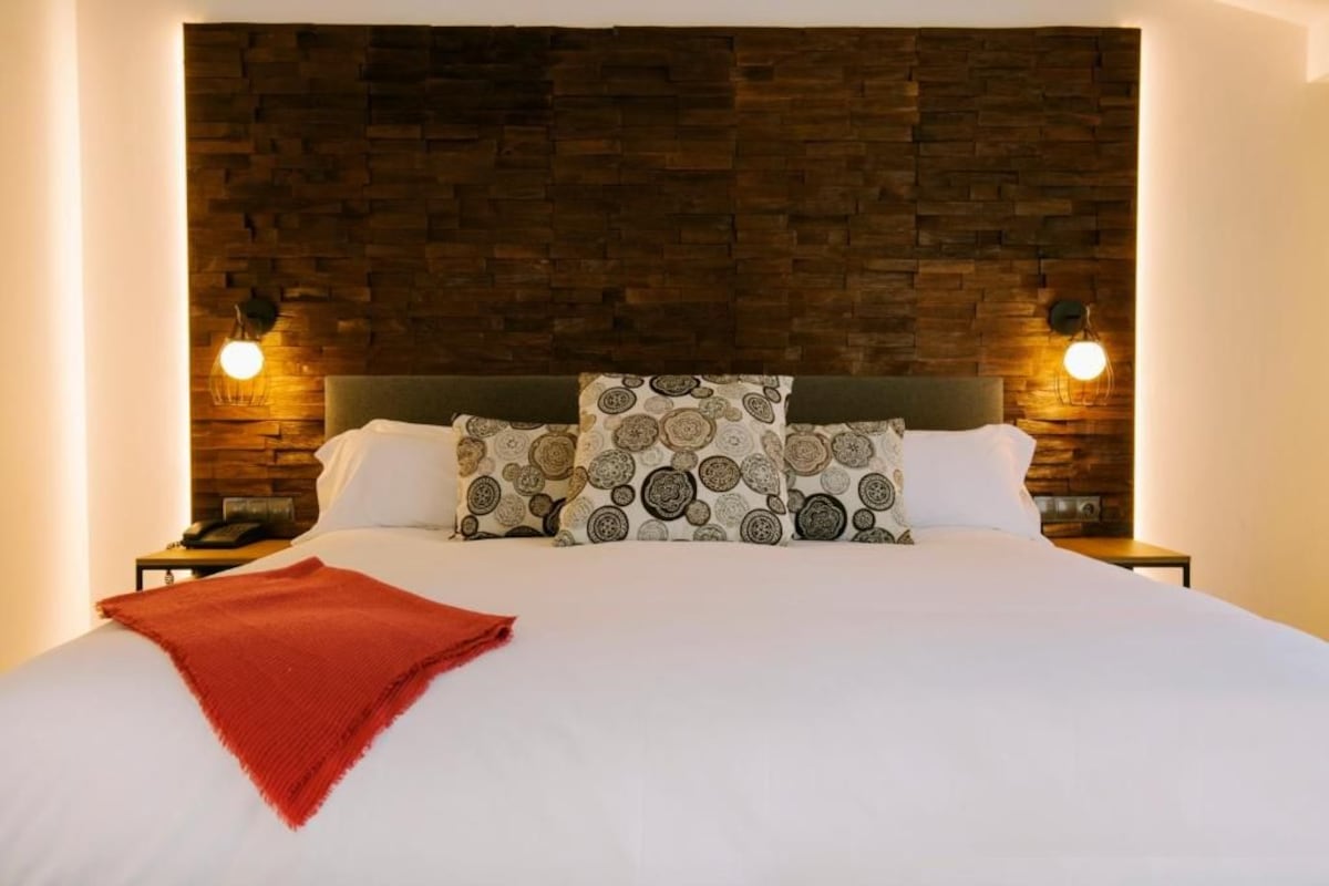 Una cama bien hecha con almohadillas estampadas, una manta roja y un cabecero de madera.