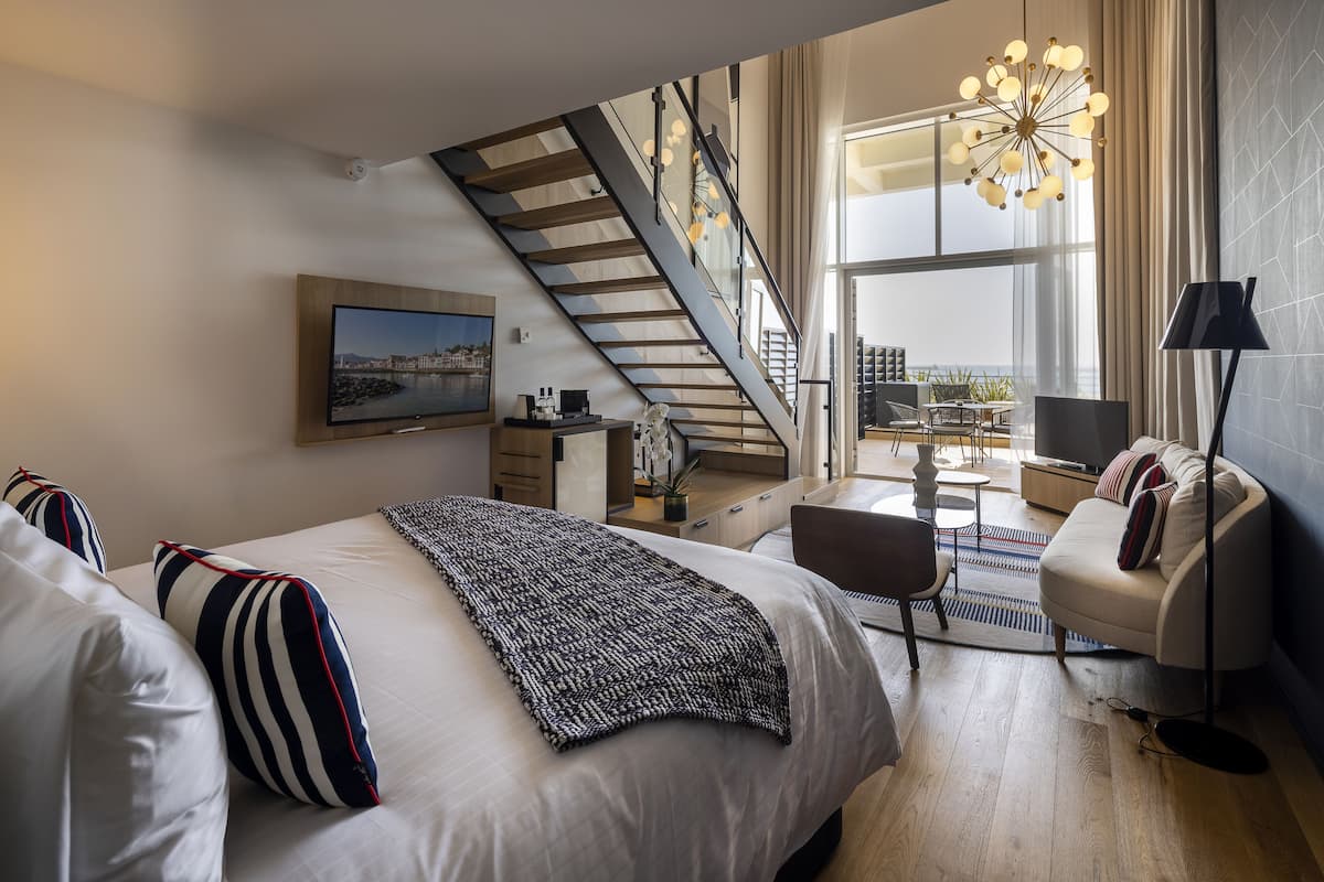 A modern bedroom with a large bed, a TV, a desk, and a staircase leading to a balcony.