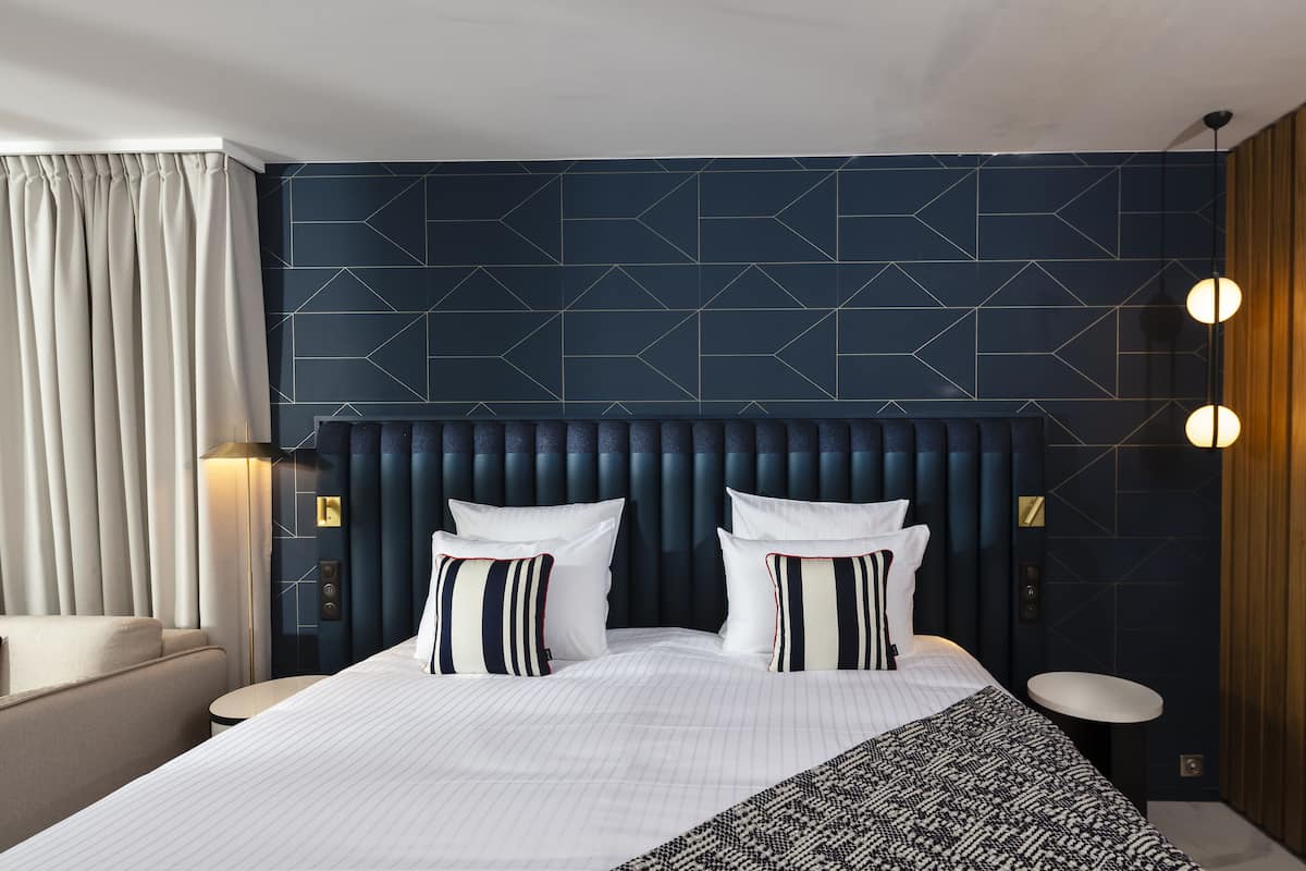 A modern hotel room with a large bed, a headboard with geometric patterns, a round table, and a wall-mounted lamp.