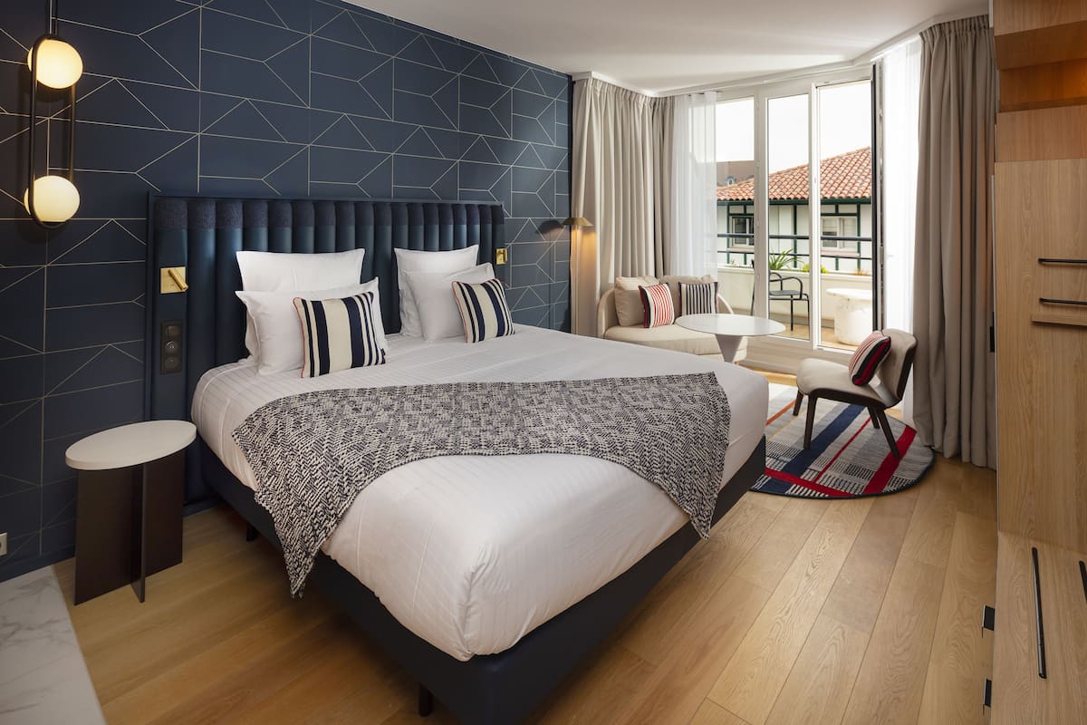 A modern hotel room with a large bed, a small round table, a sofa, and a balcony with a view.