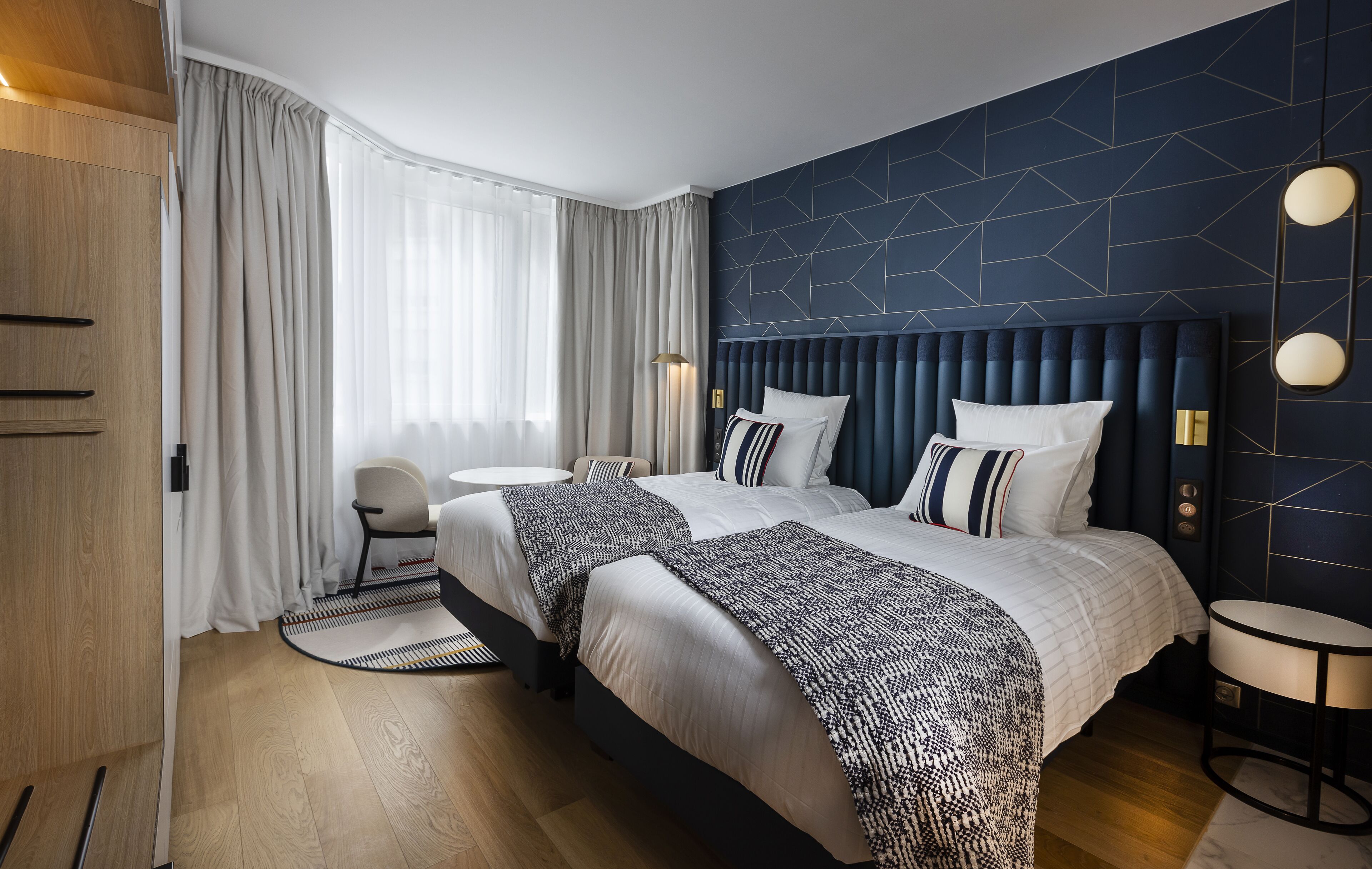 A modern hotel room with a large bed, a desk, and a chair. The room has a geometric patterned wall and a wooden wardrobe.