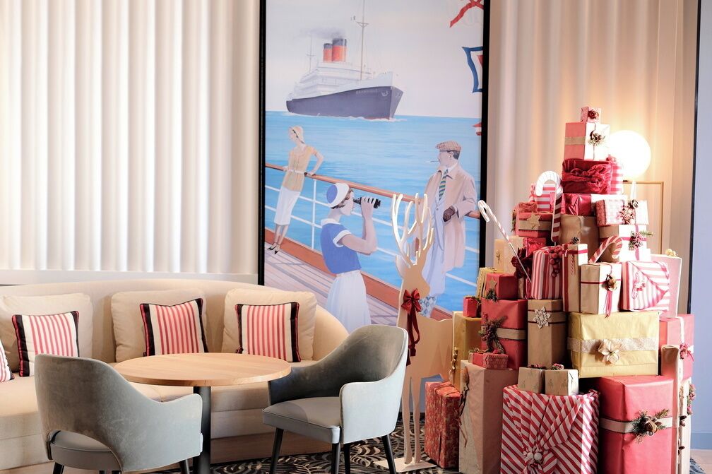 Une chambre d’hôtel moderne avec une grande fenêtre, un canapé, une table ronde et un mur orné d’une affiche représentant un navire et des personnes.