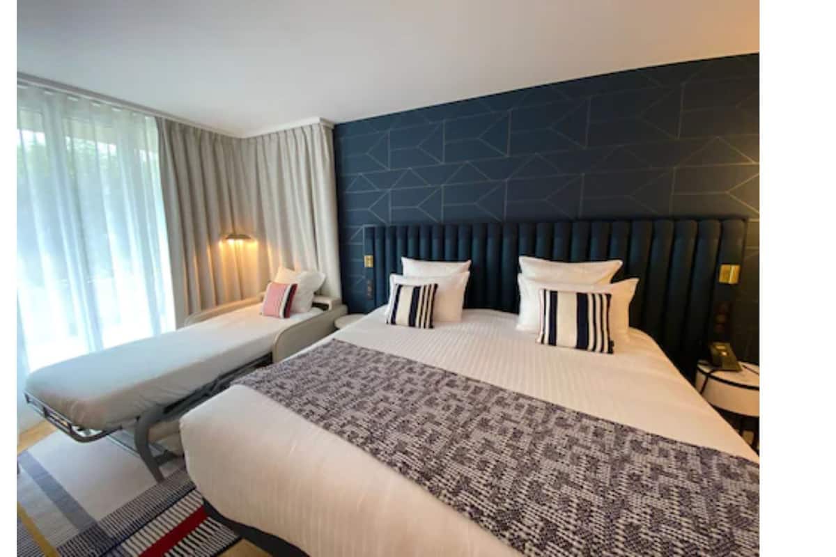 A hotel room with a bed, a bench, a large window with curtains, and a dark wall with geometric patterns.