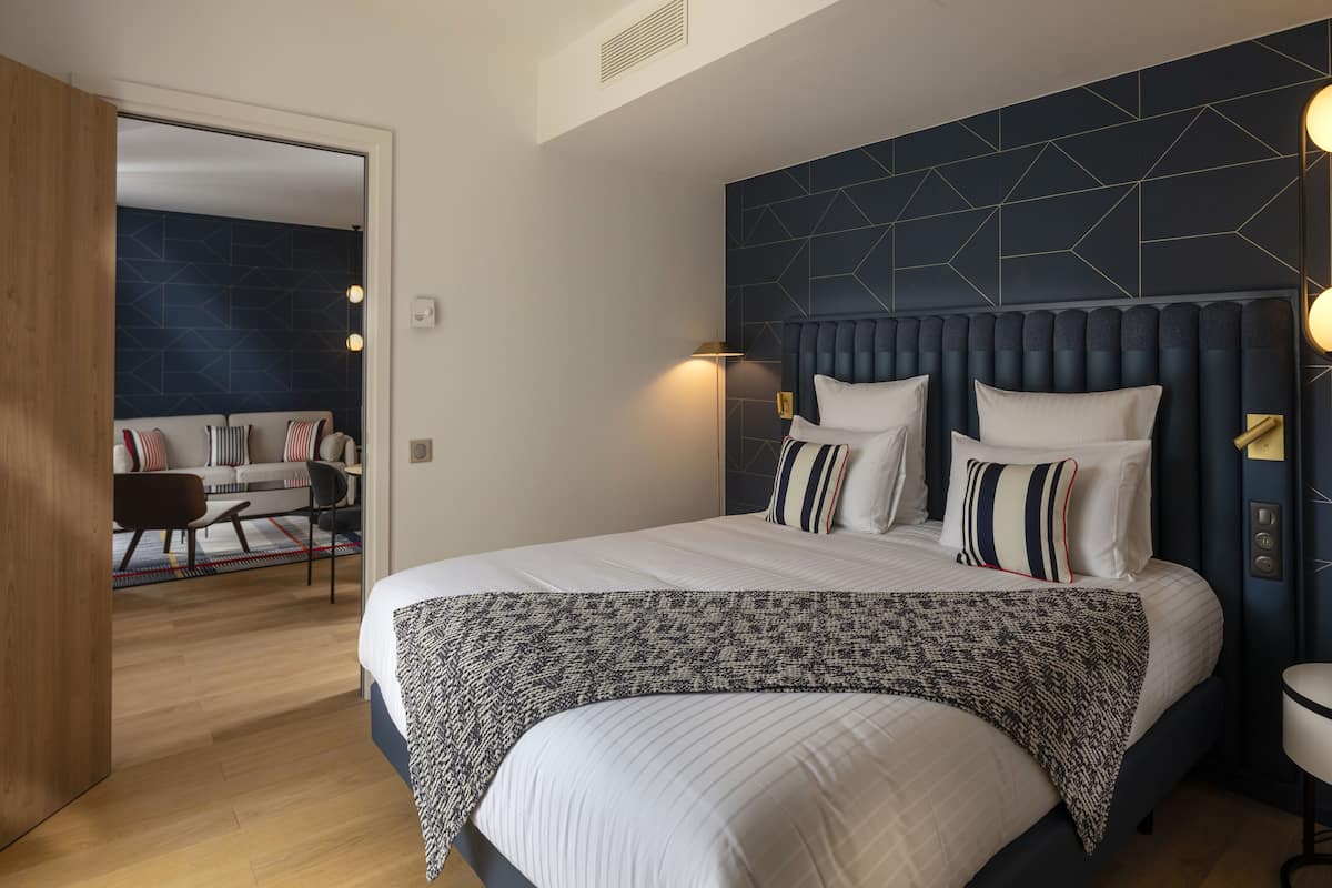 A modern bedroom with a large bed, a bedside table, and a nightstand. There is a view of a living area with a sofa and a chair.