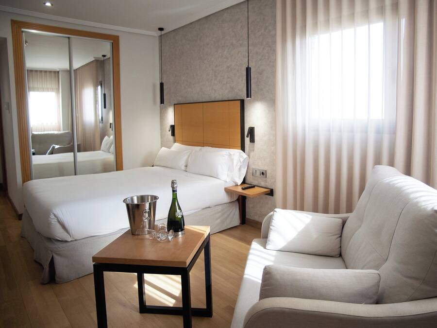 A hotel room with a large bed, a sofa, a wooden table with a bottle and glasses, and a mirror.