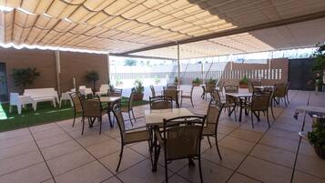 An outdoor patio area with tiled flooring, multiple tables and chairs, potted plants, and a covered section with a patterned roof.