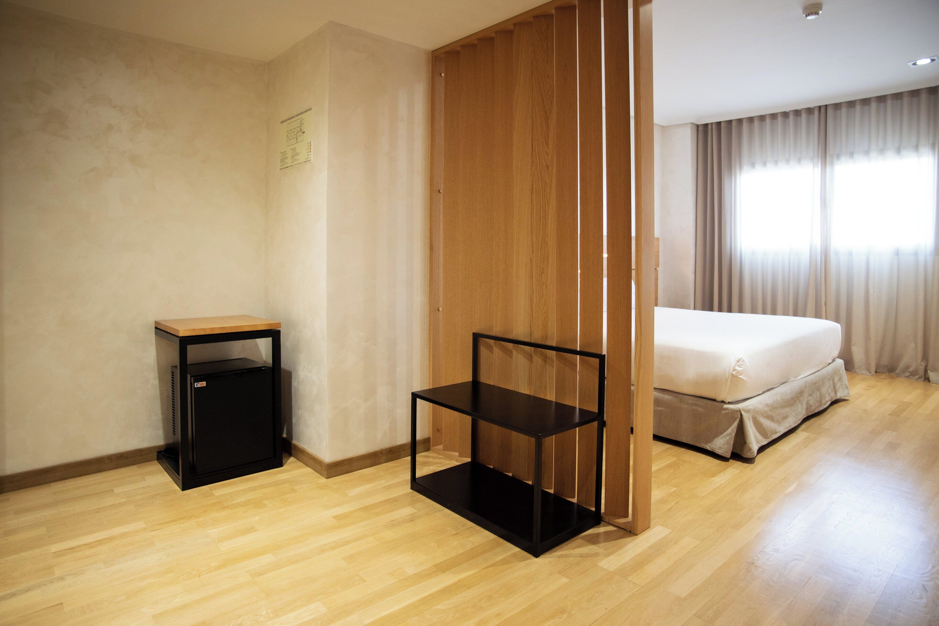 Une chambre d’hôtel avec un plancher en bois, un lit avec un matelas blanc, une table de chevet noire et une étagère noire.