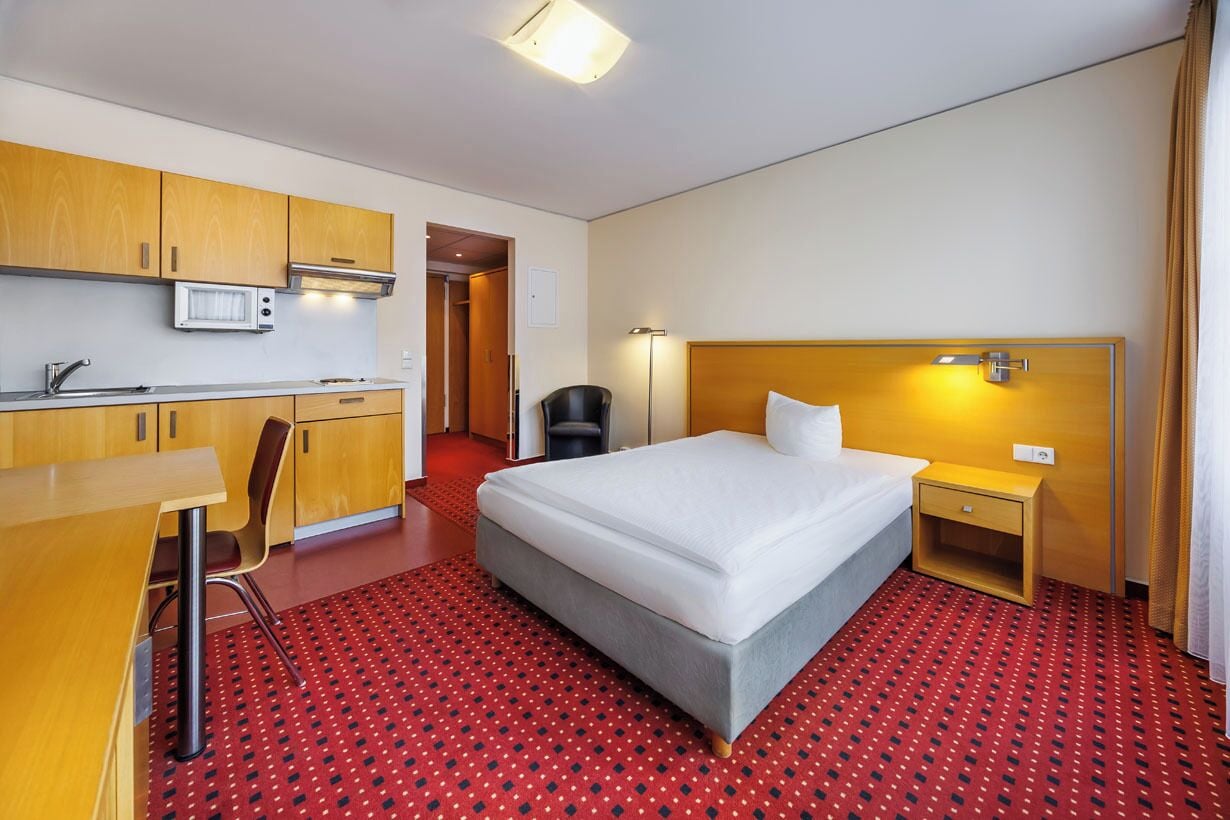 Quarto de hotel com cama, escrivaninha, cadeira, micro-ondas e pequeno espaço de cozinha.