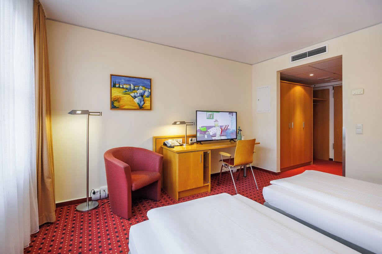 A hotel room with two beds, a desk with a computer, a chair, a TV, and a painting on the wall.