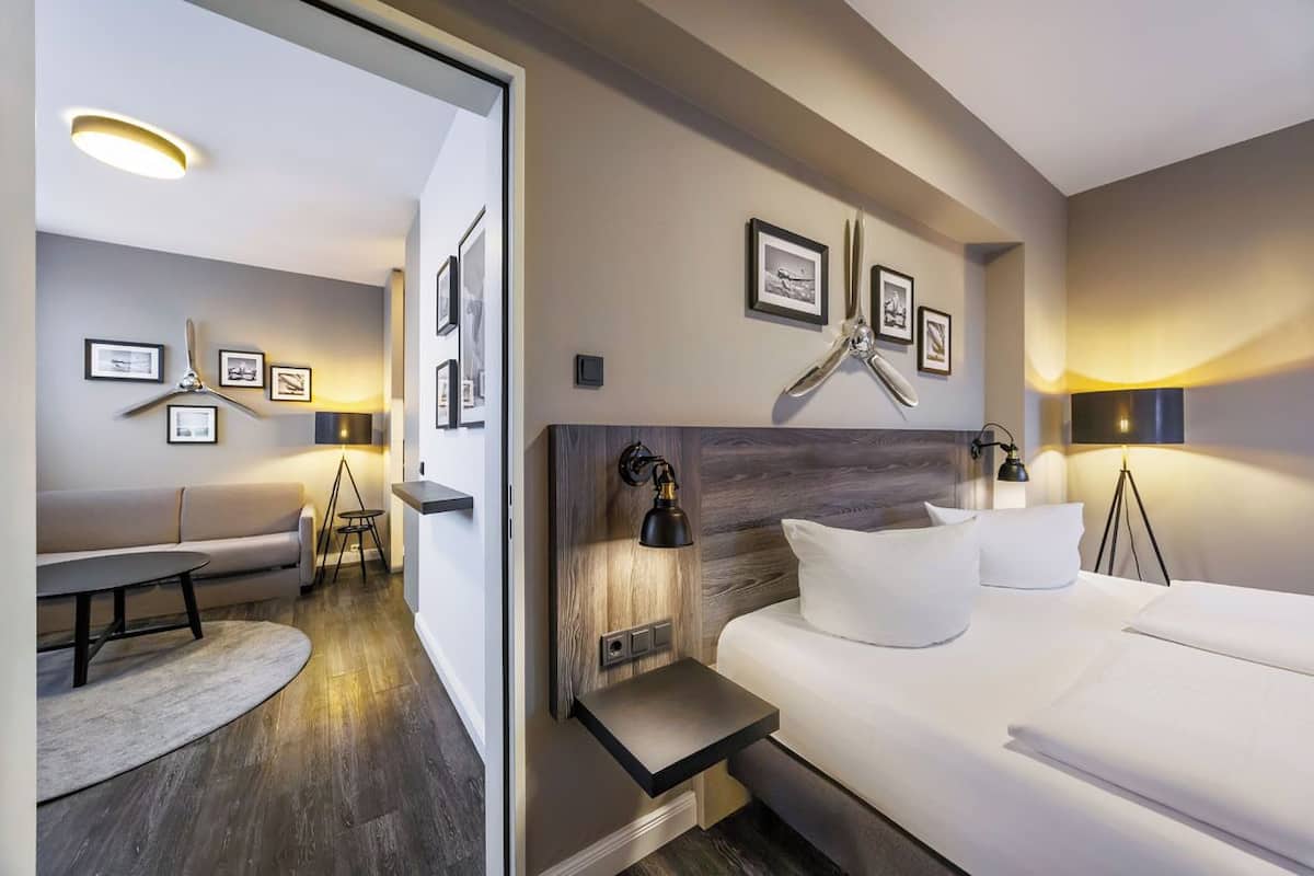 A modern hotel room with a bed, bedside table, wall-mounted lamp, and a seating area with a sofa and a small table.