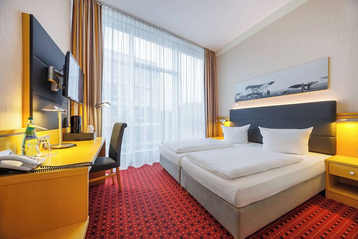 A hotel room with a bed, desk, chair, and a large window with curtains.