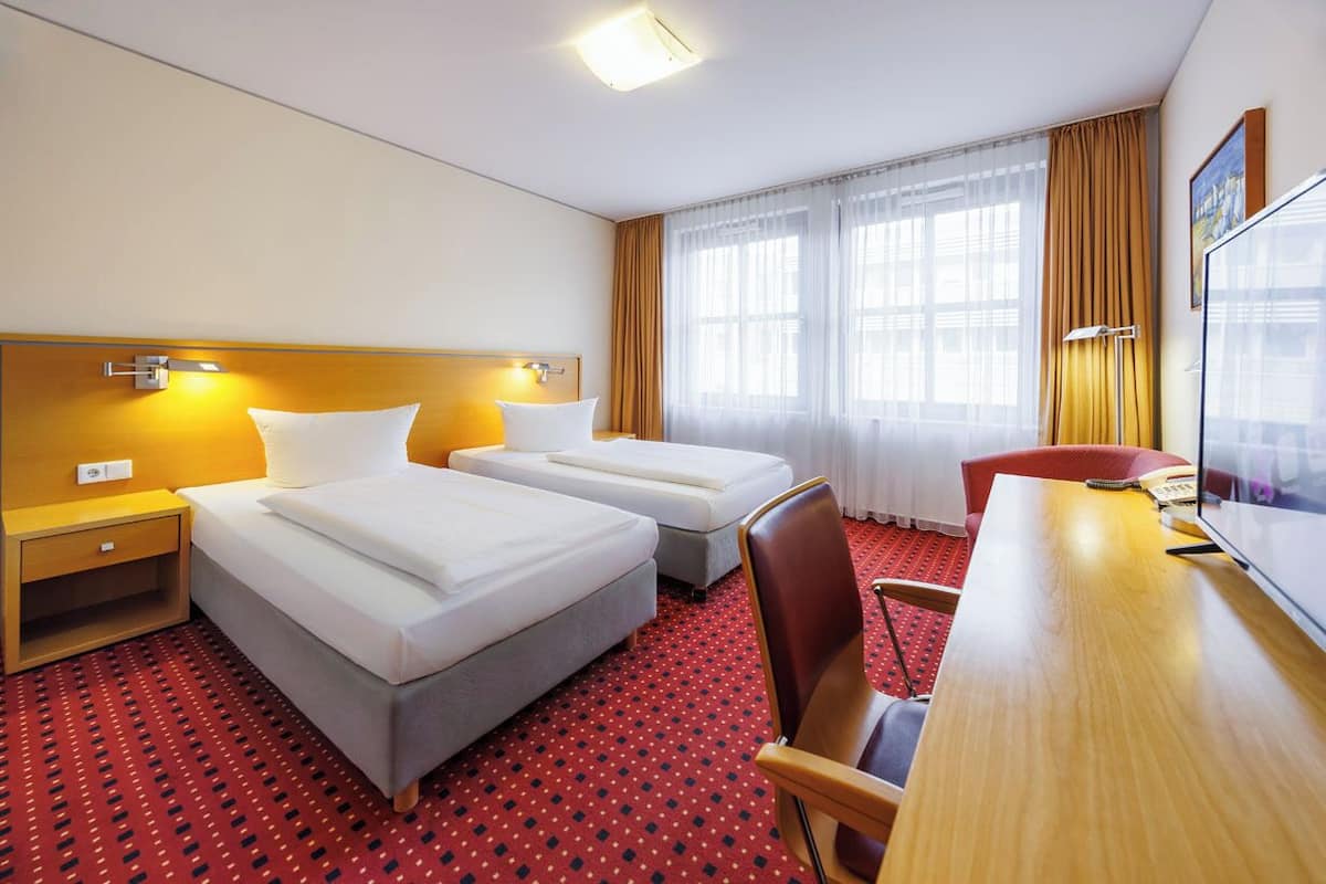 A hotel room with two beds, a desk, a chair, a TV, and a window with curtains.