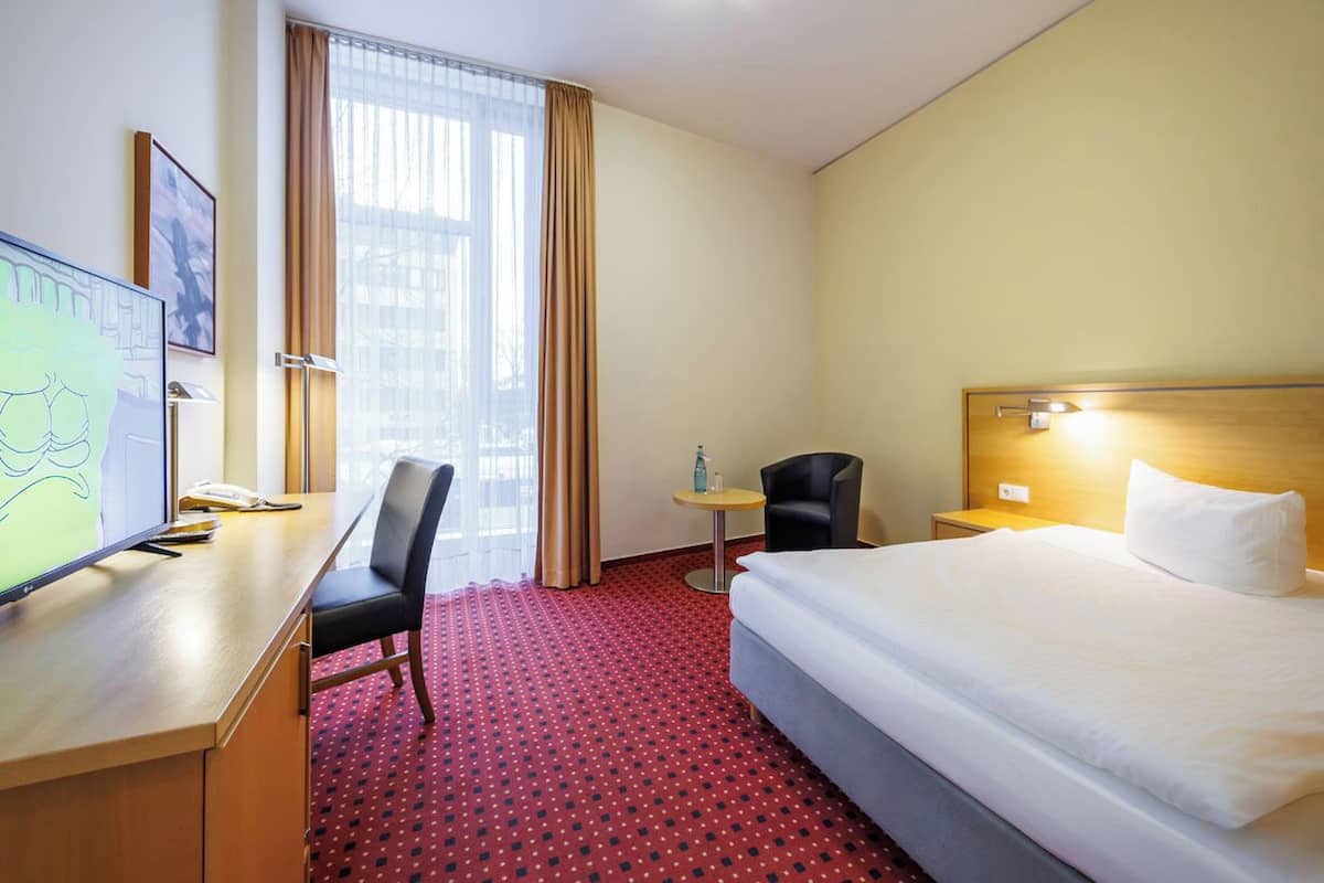 A hotel room with a bed, desk, chair, and a small table with a lamp.