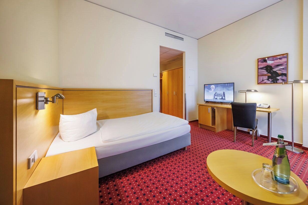 A hotel room with a bed, a desk, a chair, a TV, and a table with a bottle.