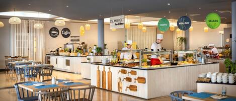 Un café moderno con mesas y sillas de madera, una barra central con exhibición de alimentos y carteles que indican café y opciones saludables.