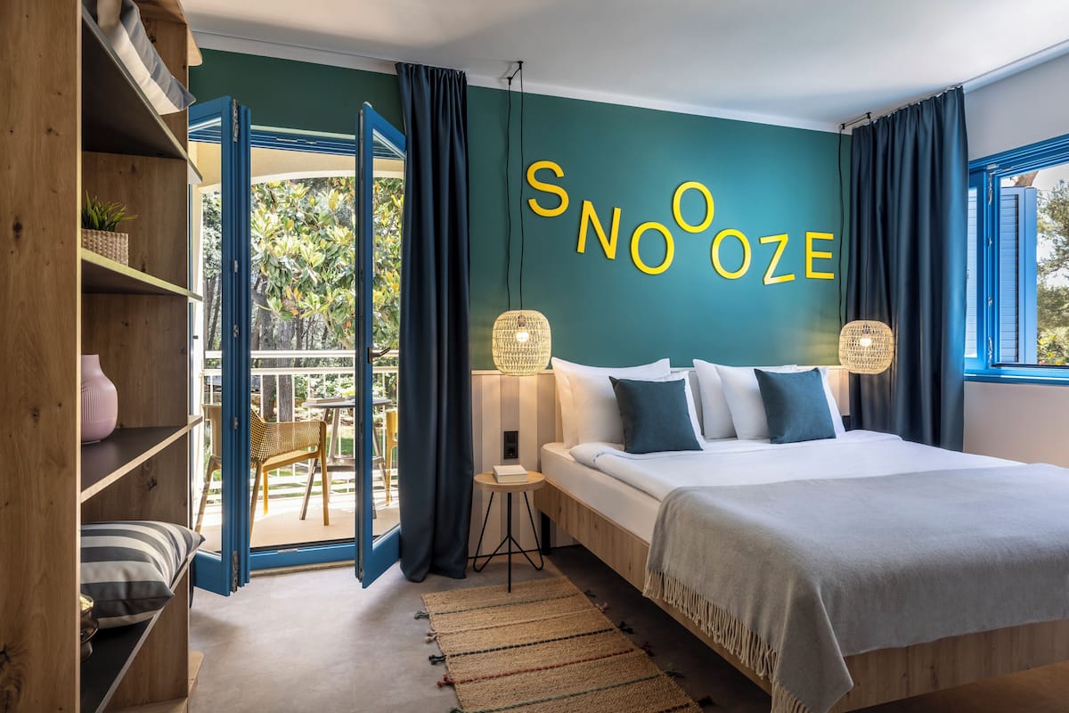 Een moderne slaapkamer met een groot bed, een wandlamp en uitzicht door een geopende deur op een buitenruimte.