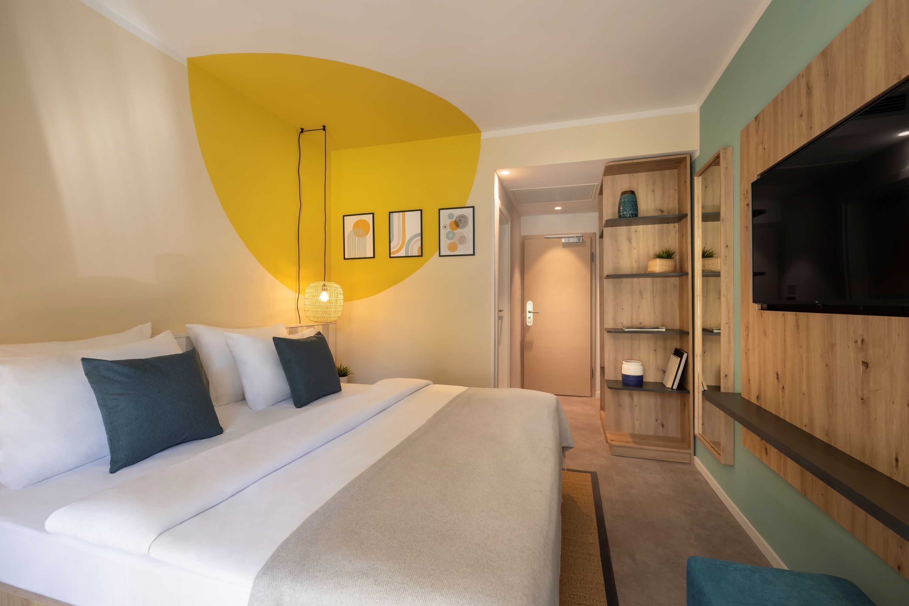 A modern hotel room with a large bed, a TV, a wooden shelf unit, and a yellow accent wall.