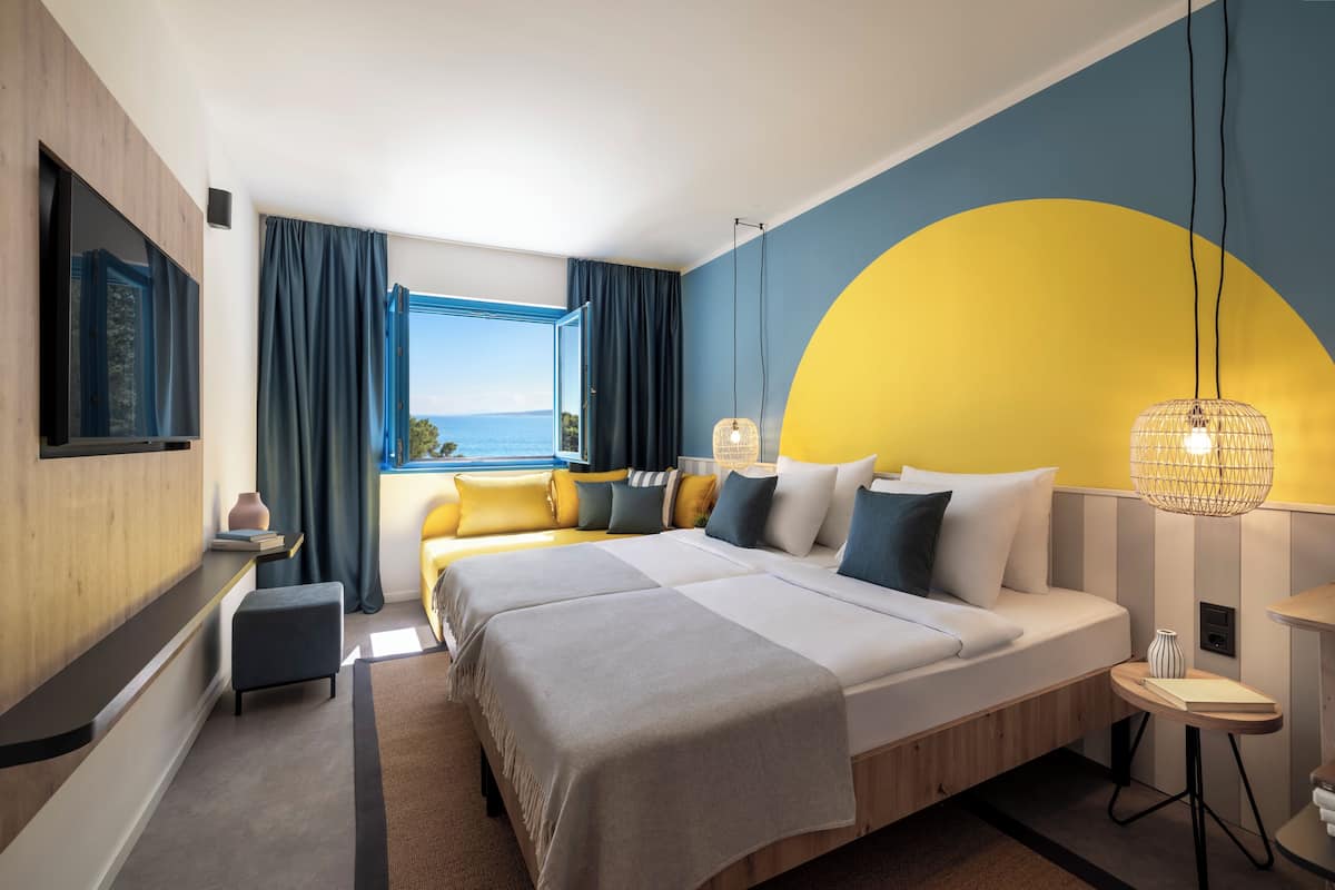 Een moderne hotelkamer met een groot bed, een televisie, uitzicht op zee en een uniek wandontwerp in geel en blauw.