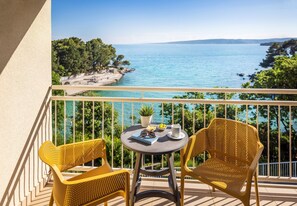 Un balcon avec deux fauteuils en osier jaune et une petite table ronde, offrant une vue sur la côte, avec une mer bleue et limpide et une végétation luxuriante.