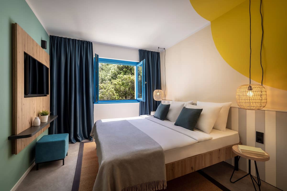 Een moderne slaapkamer met een groot bed, een televisie bevestigd aan een houten plank, een klein tafeltje en een raam met gordijnen.