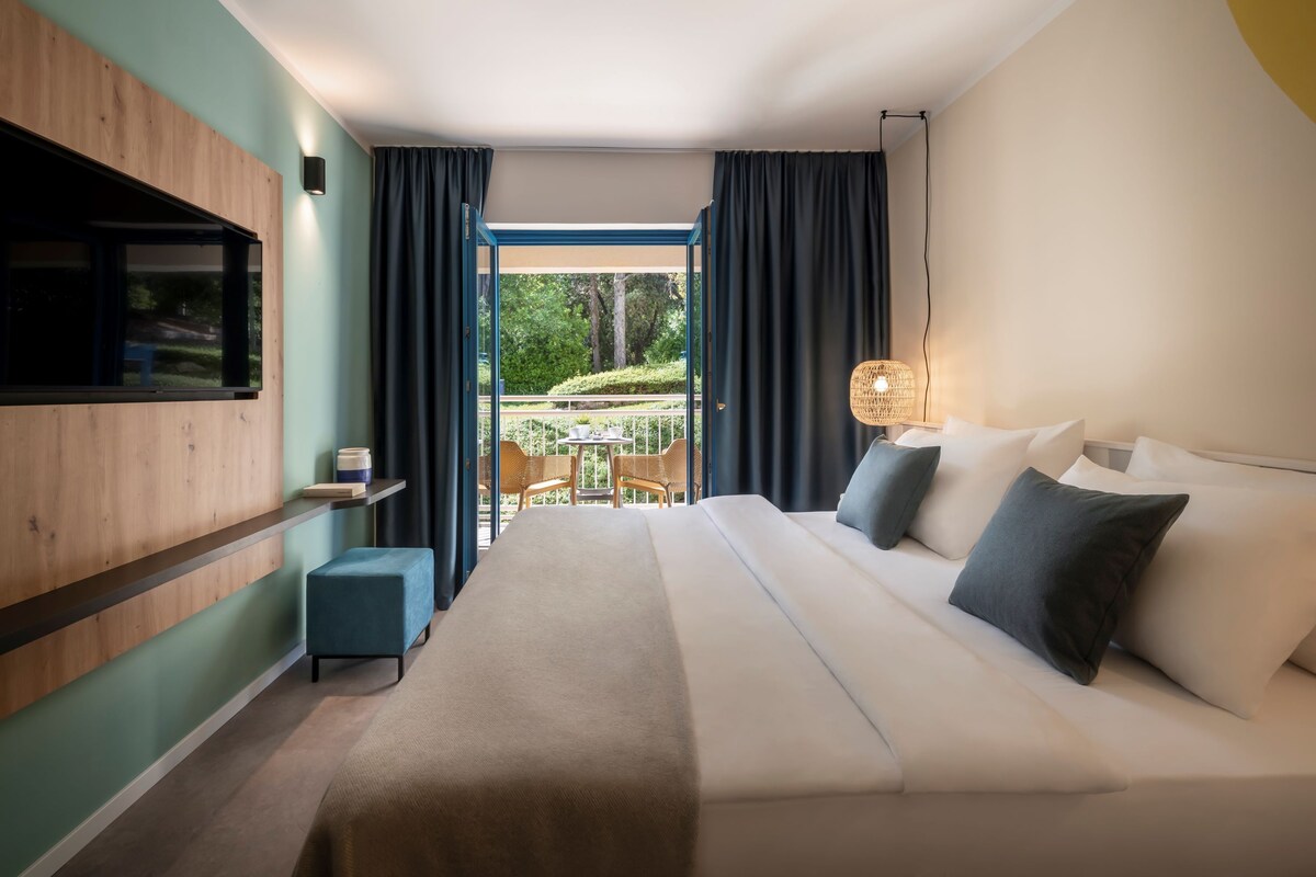 Una habitación de hotel moderna con una cama grande, un televisor montado en la pared, una pequeña zona de estar con sillas y un balcón con vistas a zonas verdes.