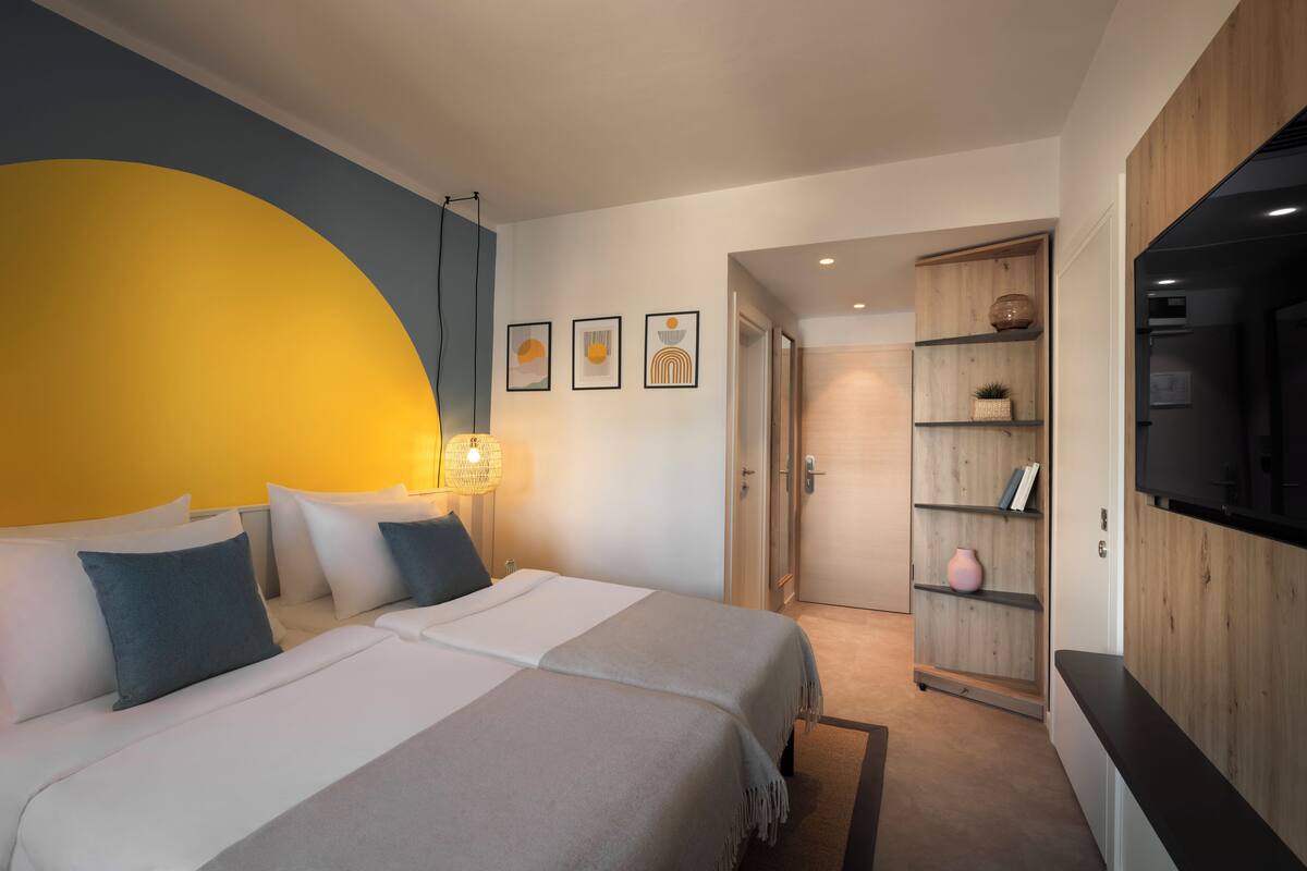 Een moderne hotelkamer met twee bedden, een grote gele accentmuur, een houten boekenplank en een televisie.