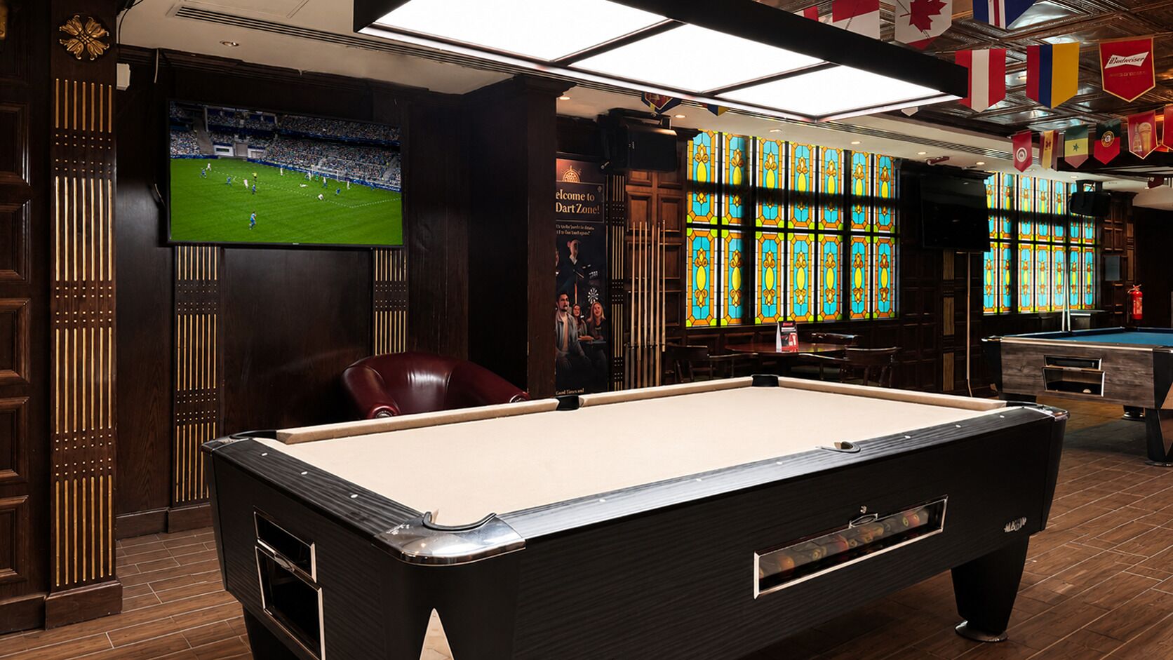 Une table de billard dans une pièce dotée d’un grand écran diffusant un événement sportif, de lambris en bois et de vitraux.