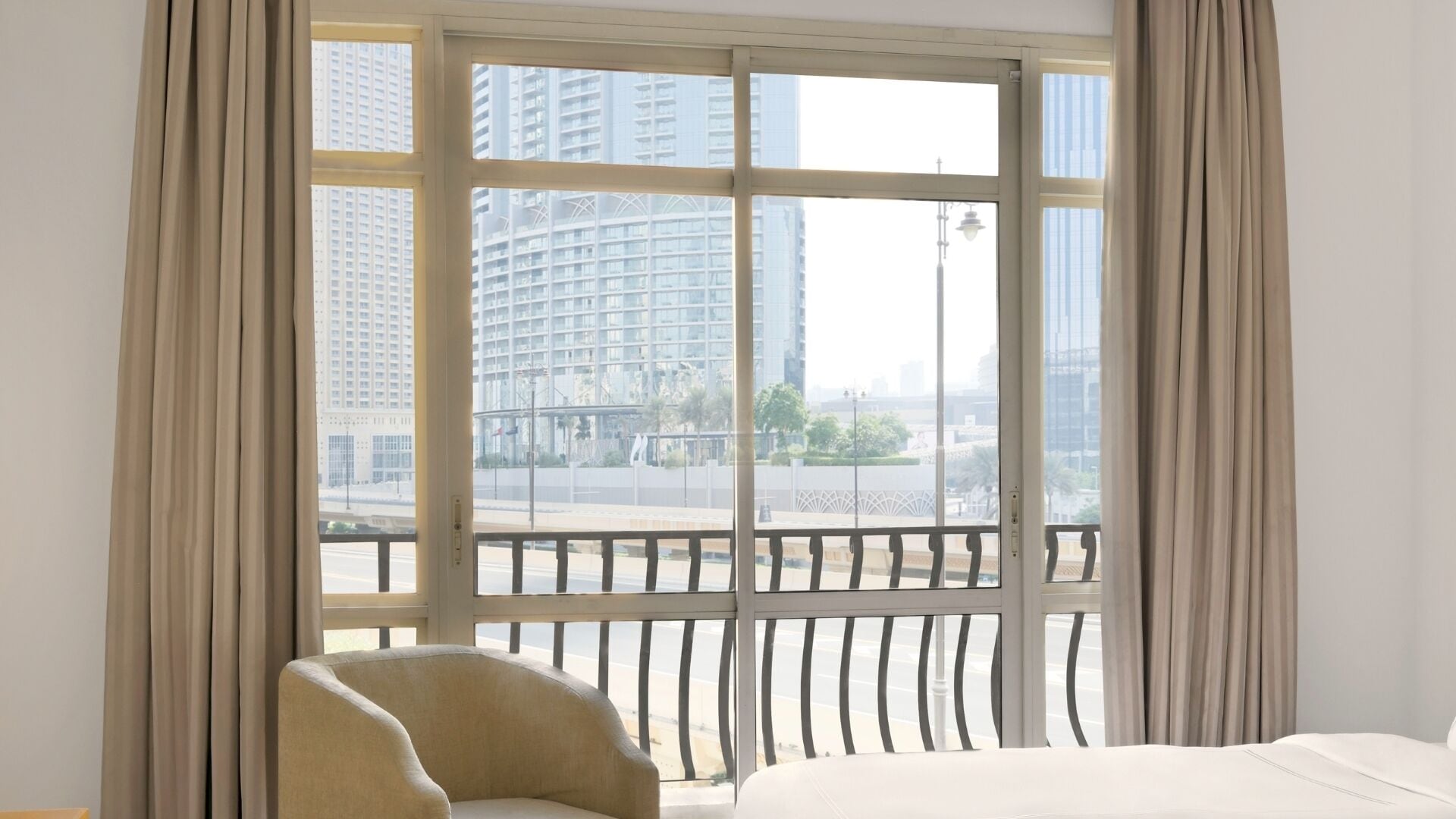 Habitación de hotel con un ventanal grande, una cama, una silla y vistas a un paisaje urbano.