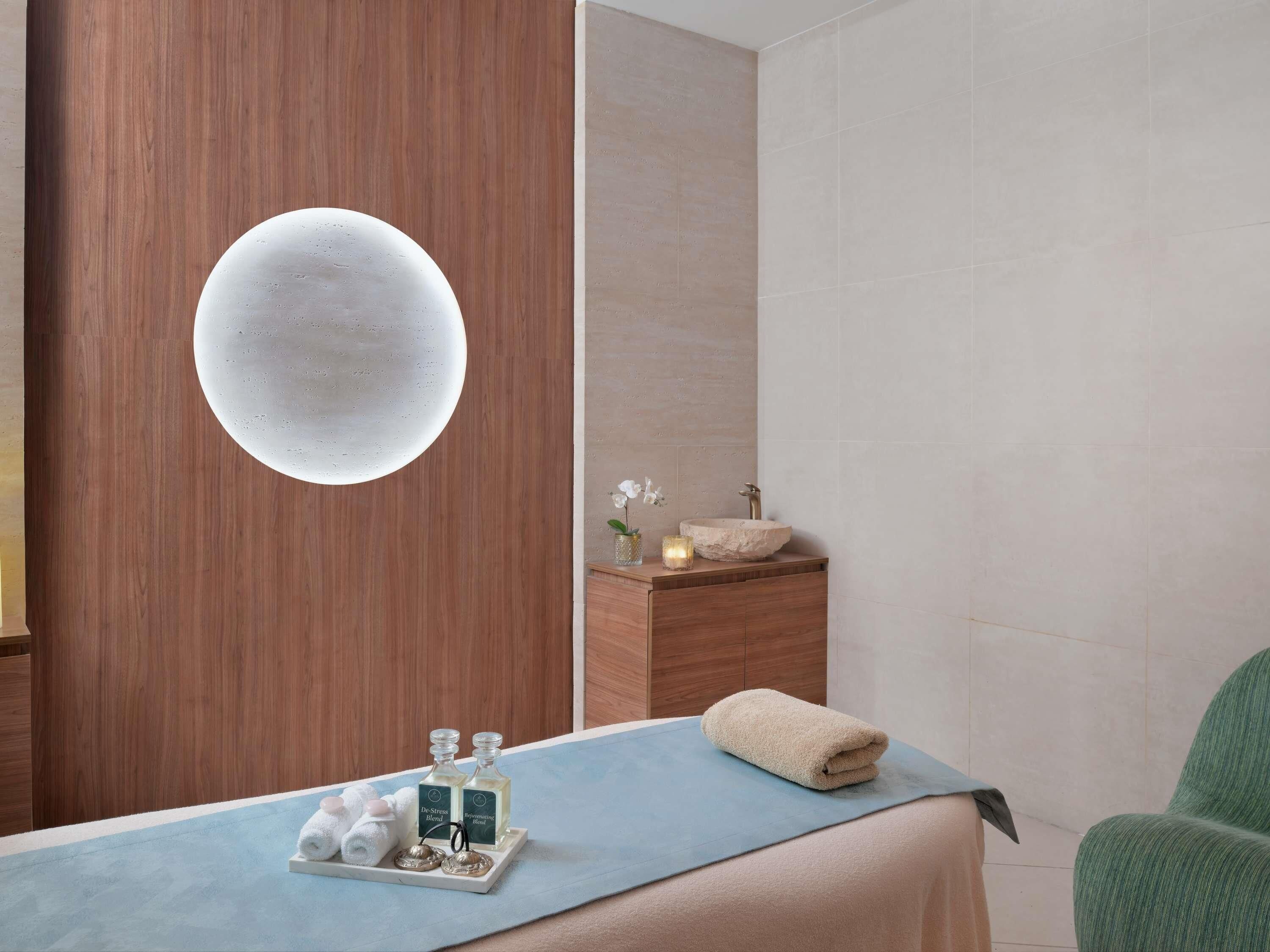 A spa room with a wooden wall, a round mirror, a sink, a towel, and a tray with bottles and towels.