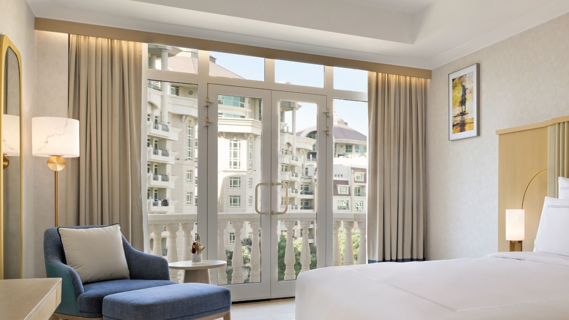 A hotel room with a bed, a chair, a small table, and a view of buildings through large windows.