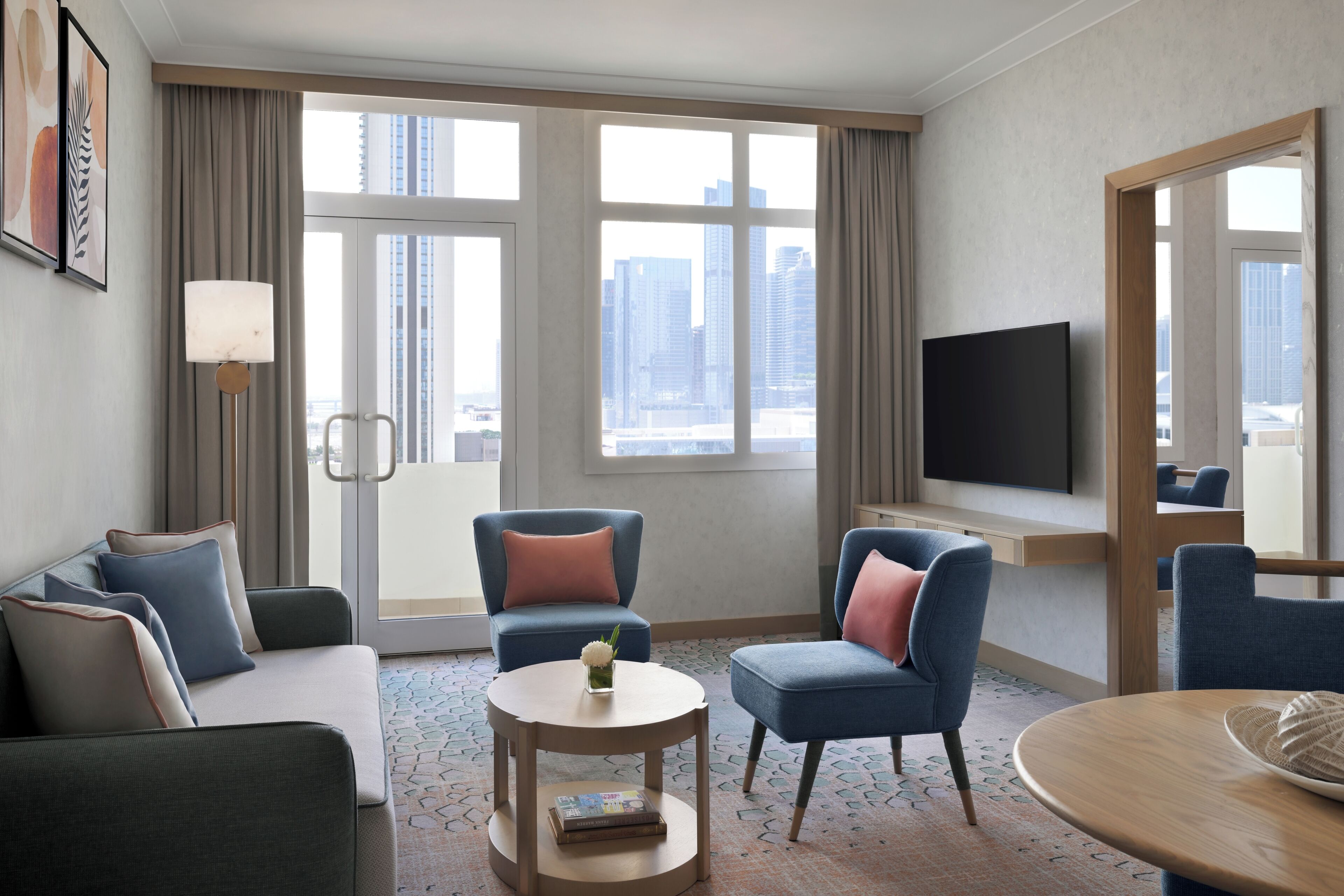 Quarto de hotel com sofá, duas poltronas, uma mesa pequena, televisão e uma janela grande com vista para a cidade.