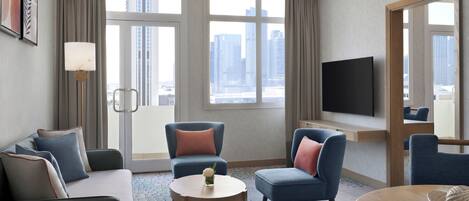 Quarto de hotel com sofá, duas poltronas, uma mesa pequena, televisão e uma janela grande com vista para a cidade.