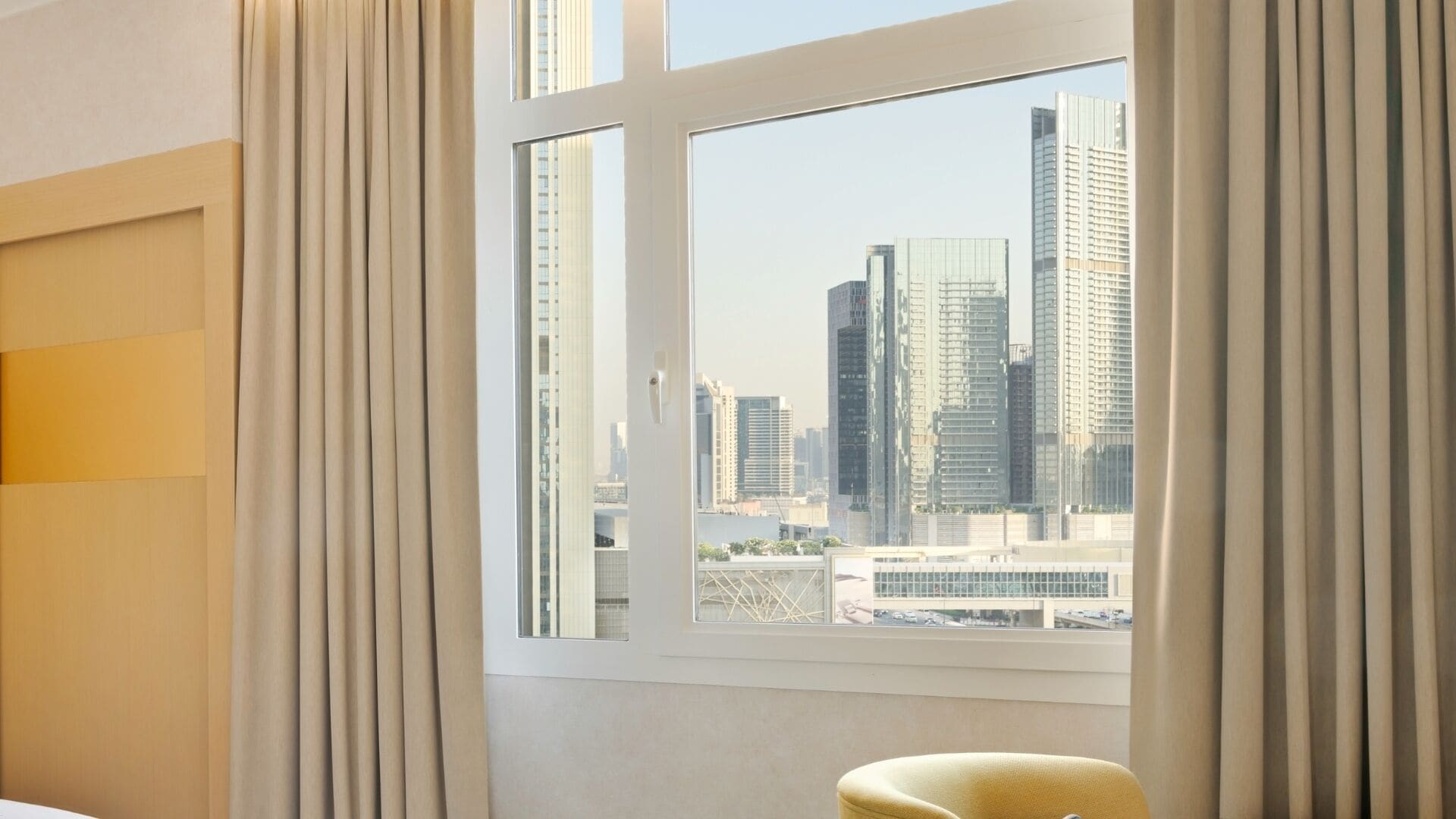 Una habitación de hotel con un ventanal grande, cortinas y vistas a edificios de la ciudad.