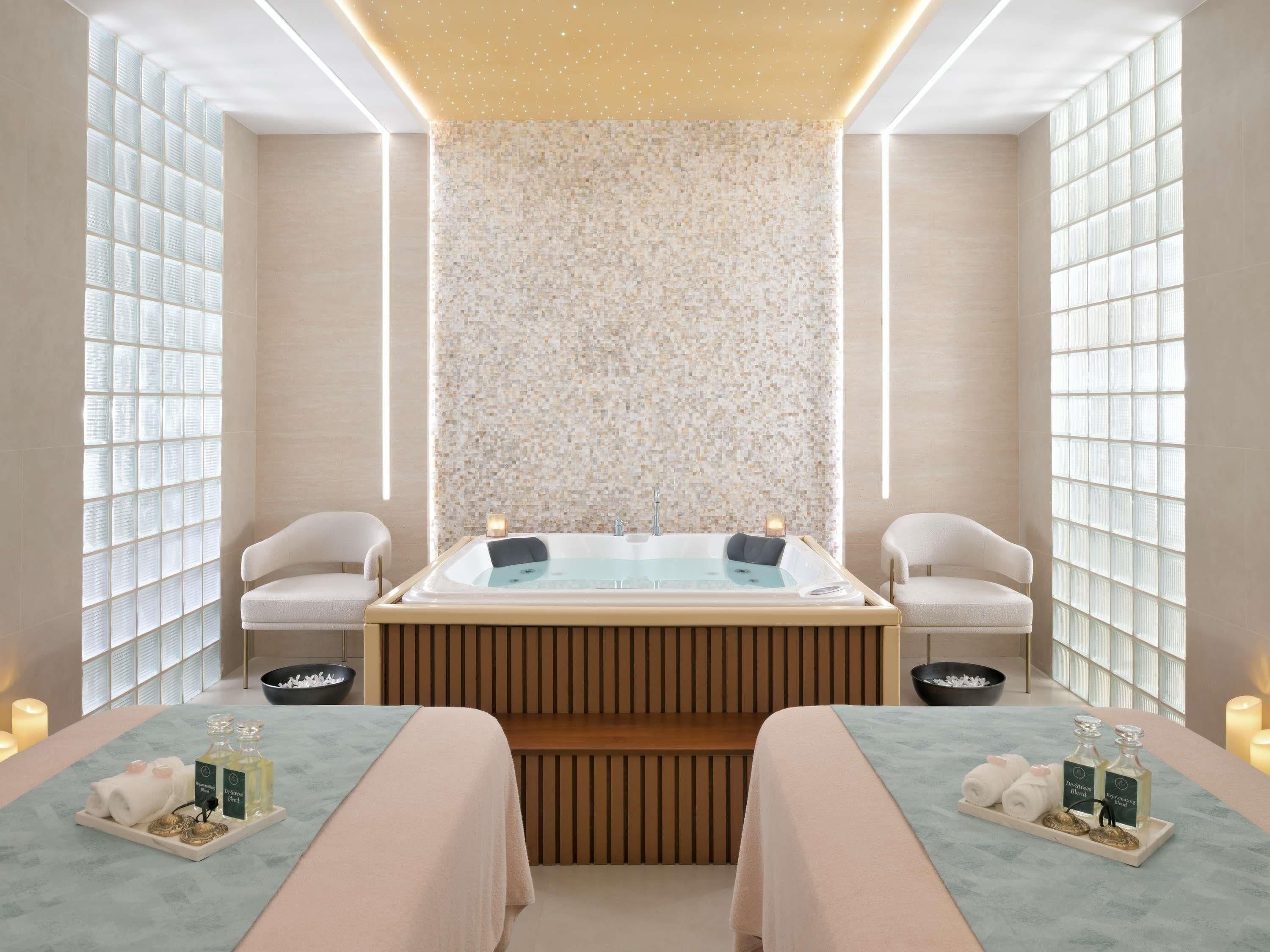 A spa room with a large bathtub, two chairs, a wooden bench, and a wall with a mosaic pattern.