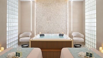 A spa room with a large bathtub, two chairs, a wooden bench, and a wall with a mosaic pattern.