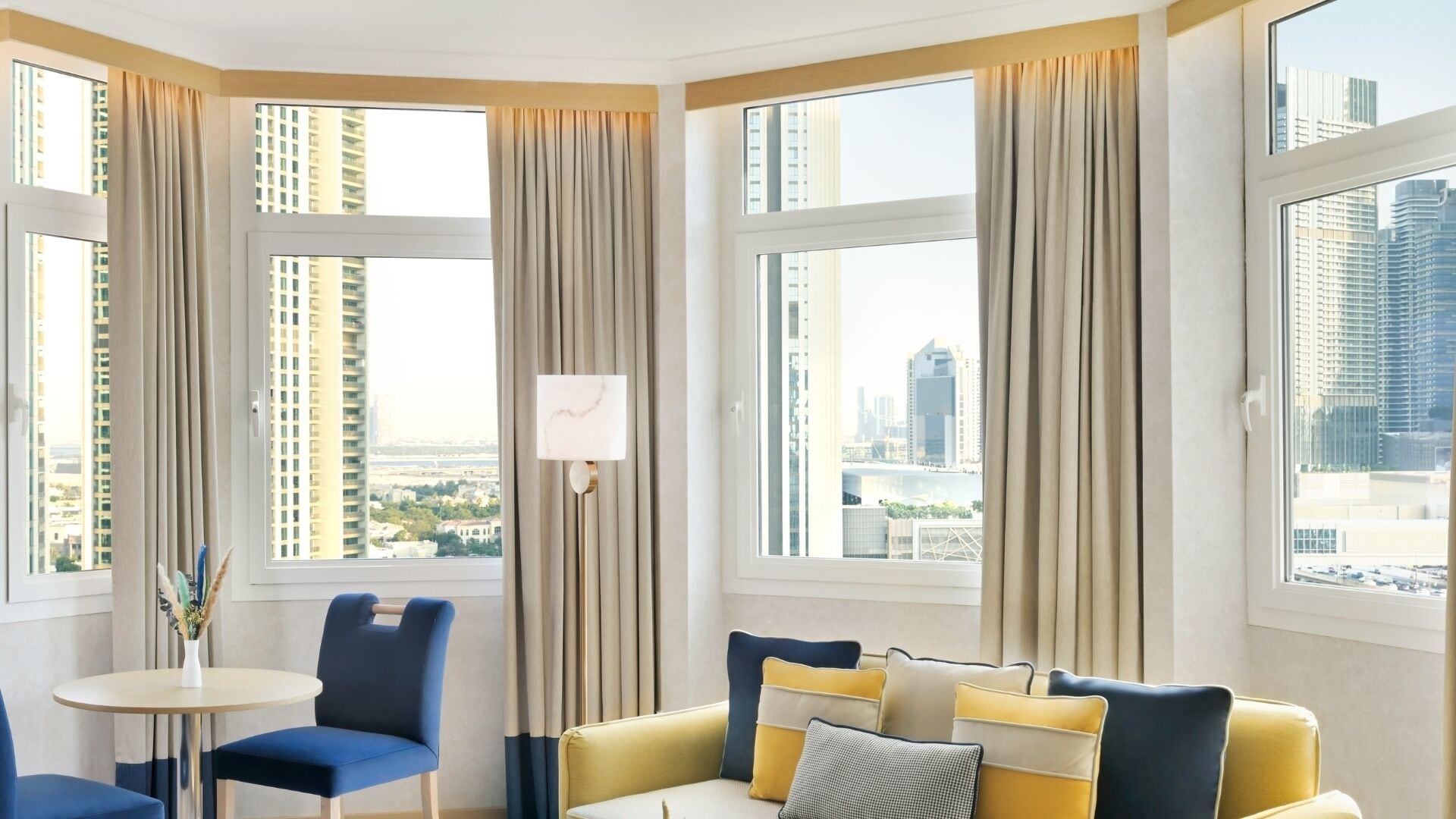 A hotel room with a city view, a sofa, a chair, and a small table with a vase.