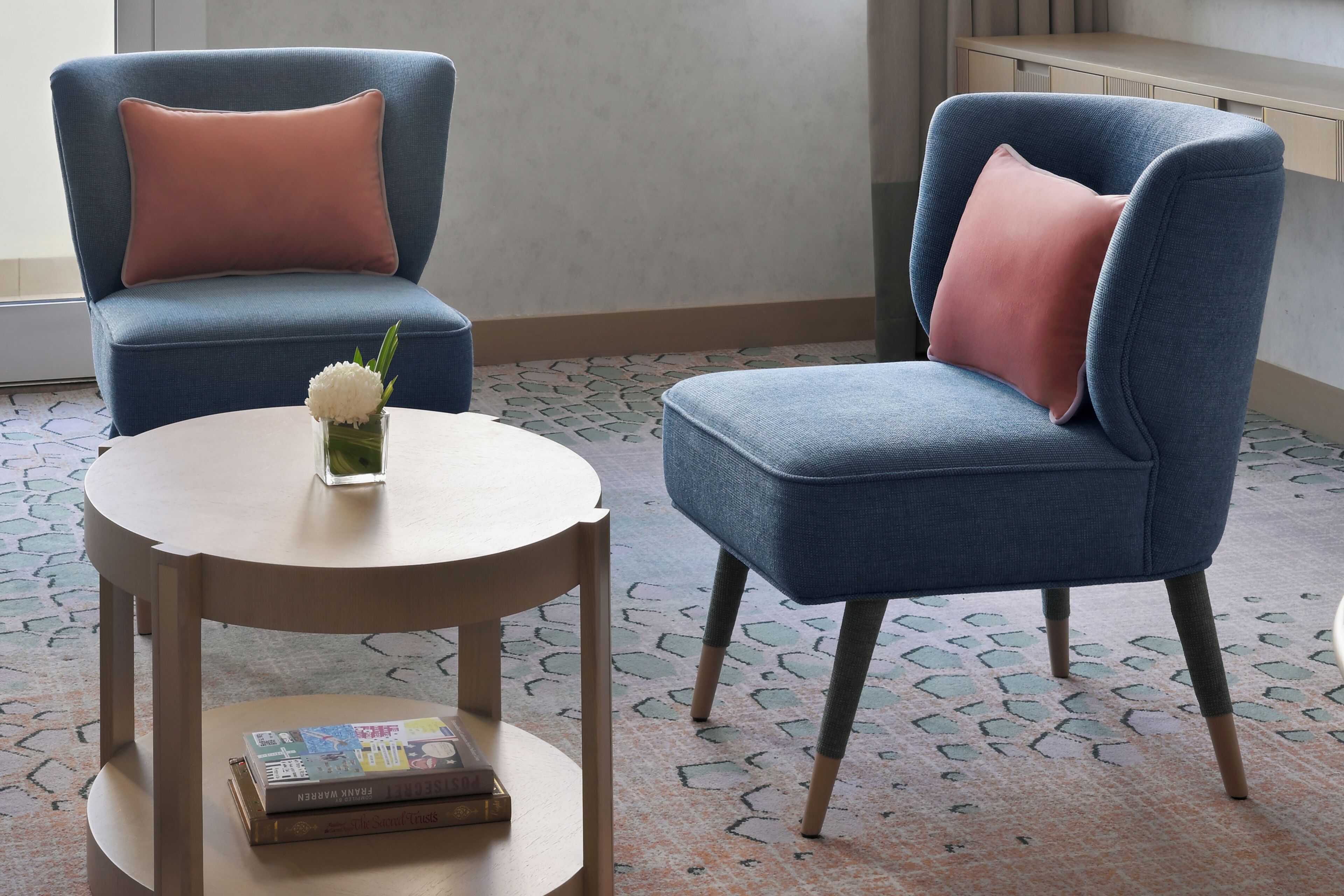Two blue armchairs with pink cushions, a round wooden table, and a vase with white flowers.