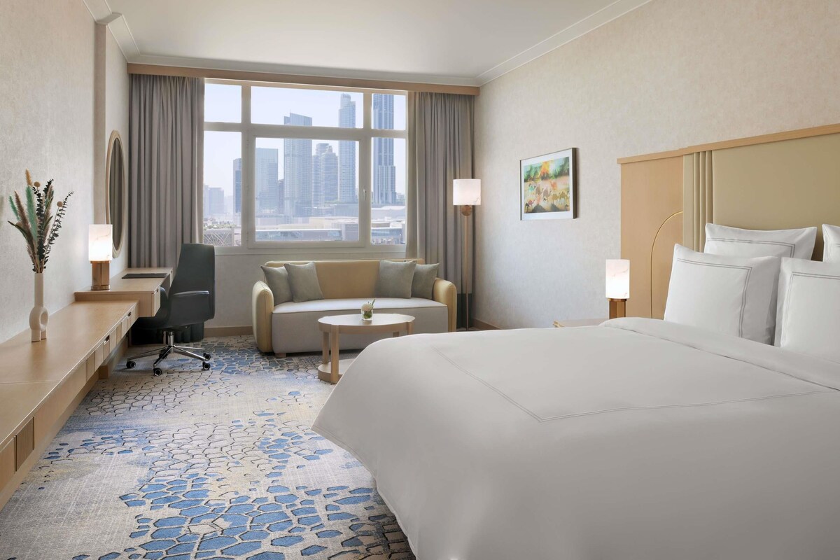 Habitación de hotel con una cama grande, un sofá, un escritorio, una silla, una mesita y vistas de la ciudad desde la ventana.