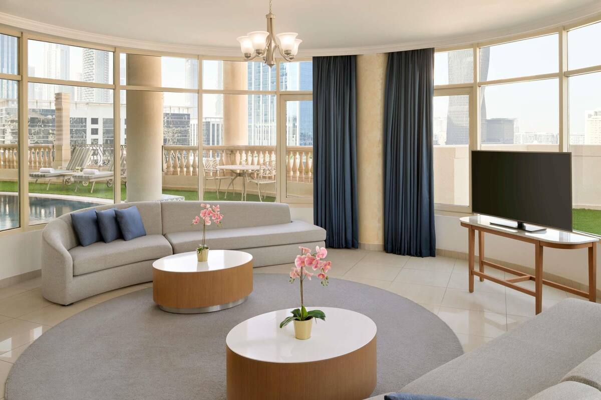 A modern living room with a large sofa, two round coffee tables, a TV on a stand, and a view of a cityscape through large windows.