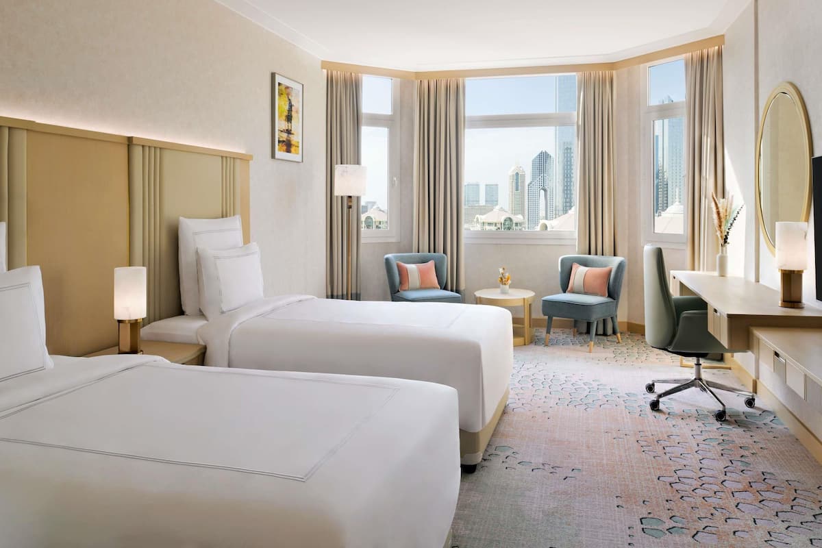 A hotel room with two beds, a desk, a chair, a small table, and a view of the cityscape.