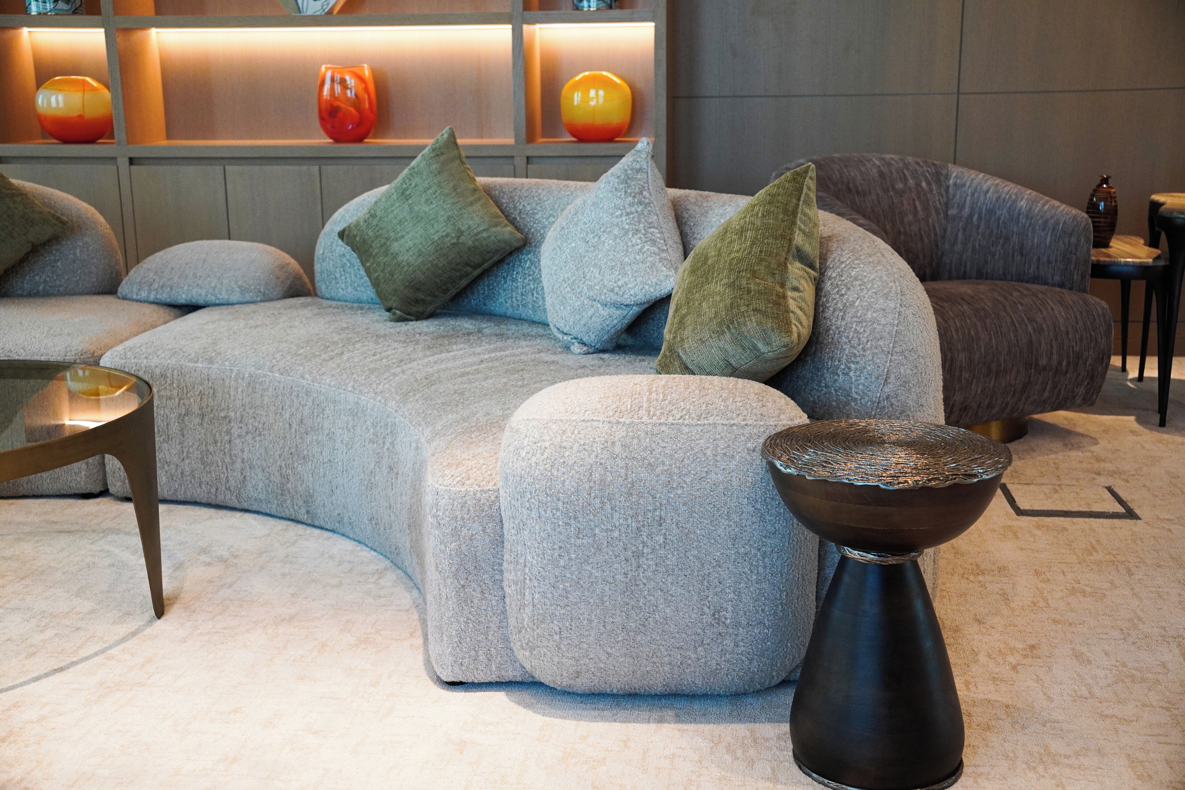 Una sala moderna con un sofá seccional gris, varios cojines, una mesita redonda y jarrones decorativos en una estantería.