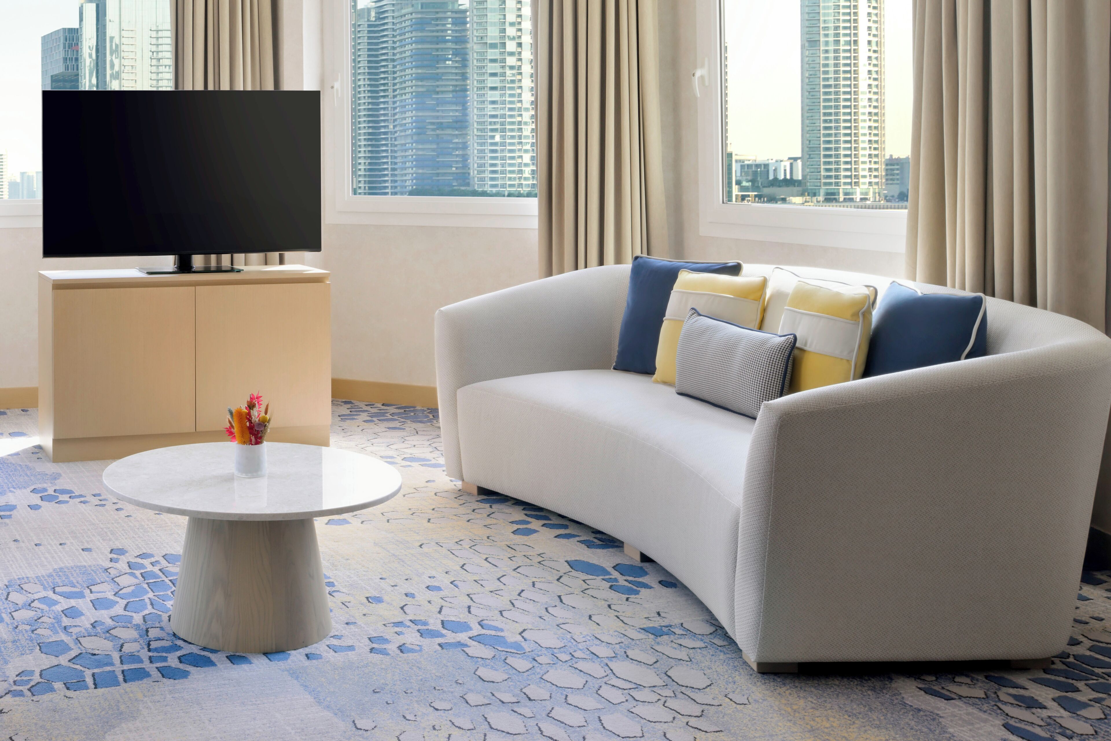 Ein modernes Wohnzimmer mit Flachbildfernseher, einer Couch mit Kissen, einem kleinen Tisch und einem gemusterten Teppich.