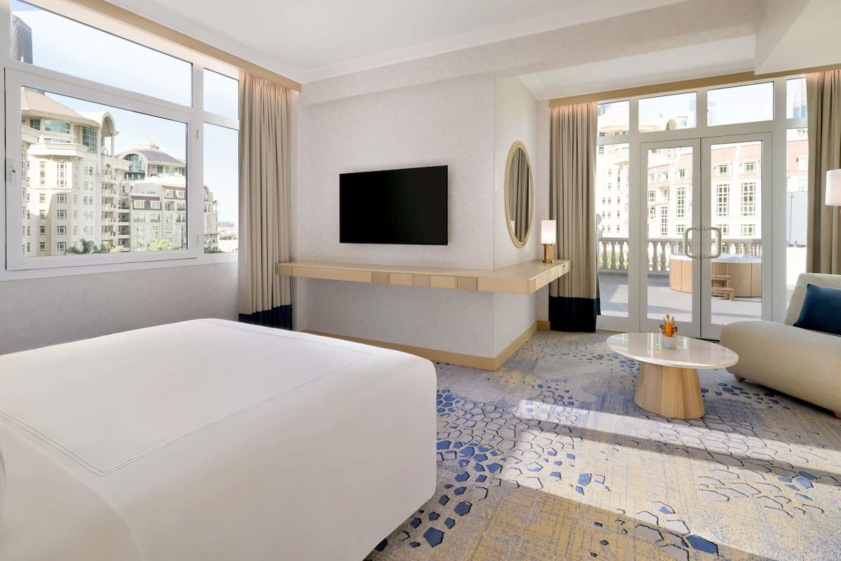 A hotel room with a bed, a TV, a small table, and a balcony with a view of buildings.