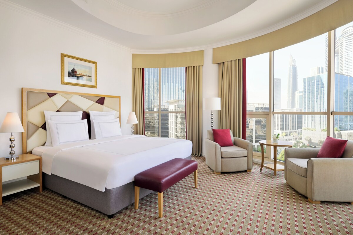 Une chambre d’hôtel avec un grand lit, deux fauteuils, une petite table et un banc.