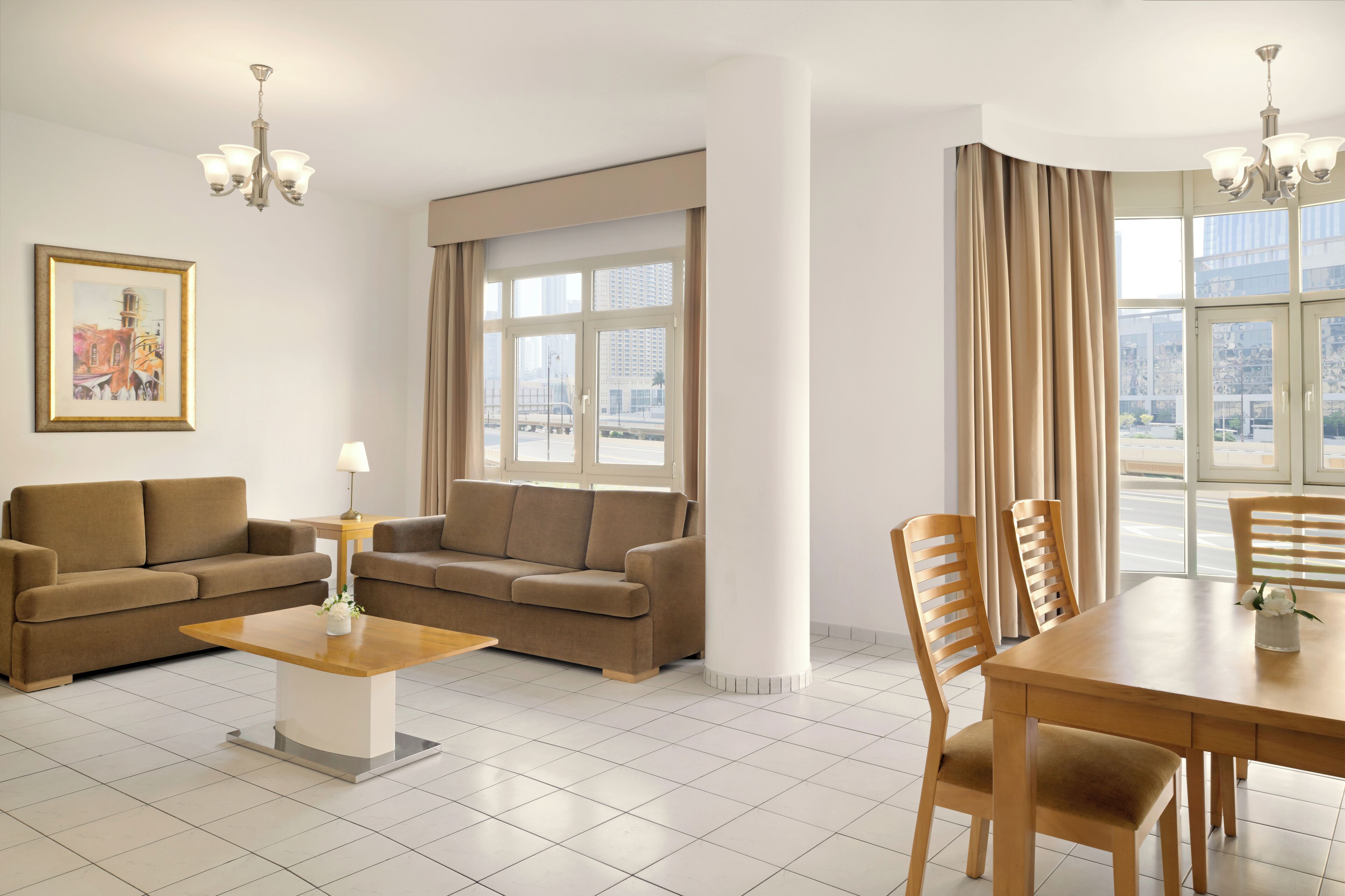 Una sala de estar moderna con dos sofás, una mesa de centro y una mesa de comedor con sillas. Hay ventanas grandes con cortinas, un candelabro y un cuadro enmarcado en la pared.