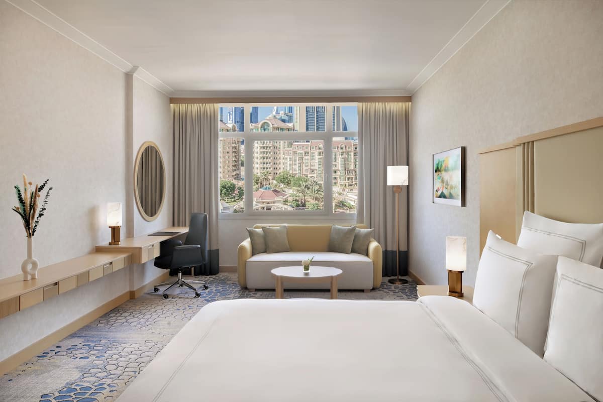 A hotel room with a large bed, a sofa, a desk, a chair, a small table, and a window with a city view.