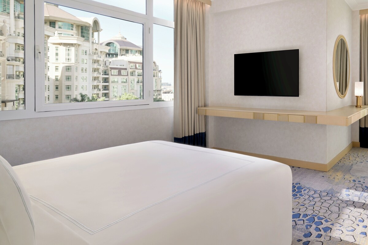 Une chambre d’hôtel avec un lit, un téléviseur fixé au mur, une fenêtre donnant sur des immeubles et un sol carrelé.