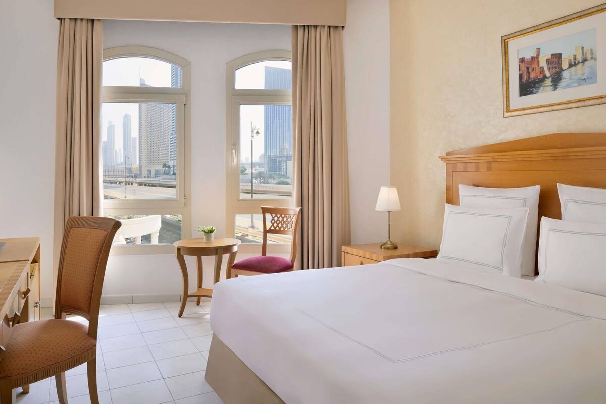 A hotel room with a large bed, a desk, a chair, a small table with a plant, and a view of a cityscape through two windows.