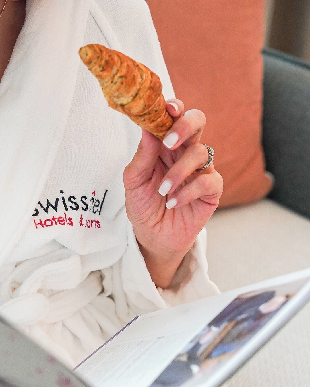 Henkilö istuu sohvalla, jossa on tyyny, ja hänellä on kädessään croissant. Hänellä on yllään valkoinen kaapu, jossa on tekstiä.