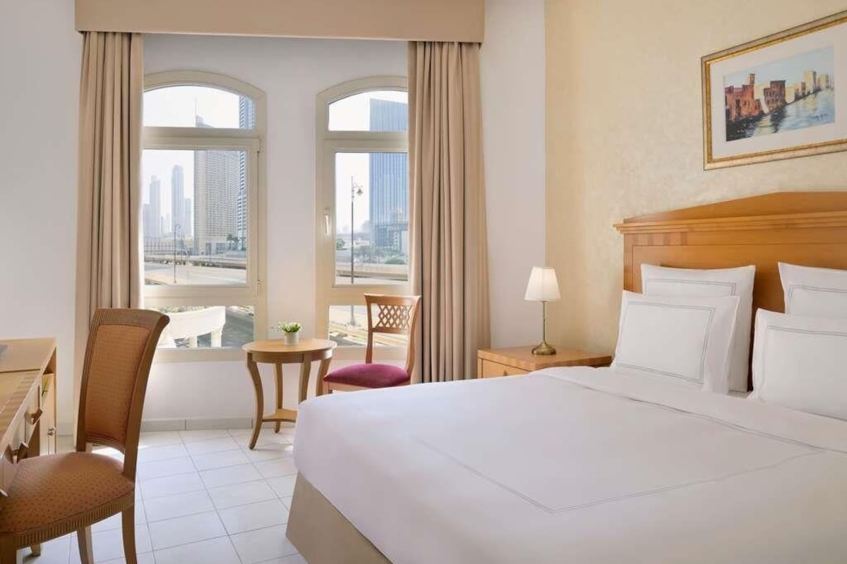 Une chambre d’hôtel avec un grand lit, un bureau, une chaise, une petite table et une vue sur le paysage urbain, visible à travers deux fenêtres.